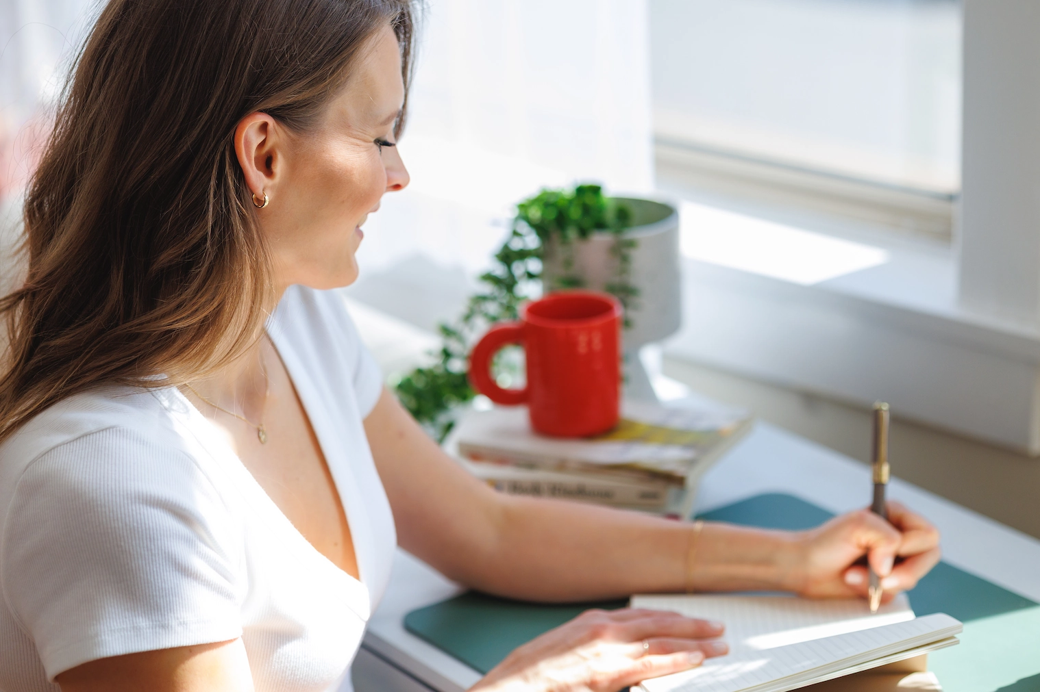 Woman sits at a desk by a window, writing in a notebook. Vegan dietitian books, a red mug, and a potted plant fill the sunlit space.