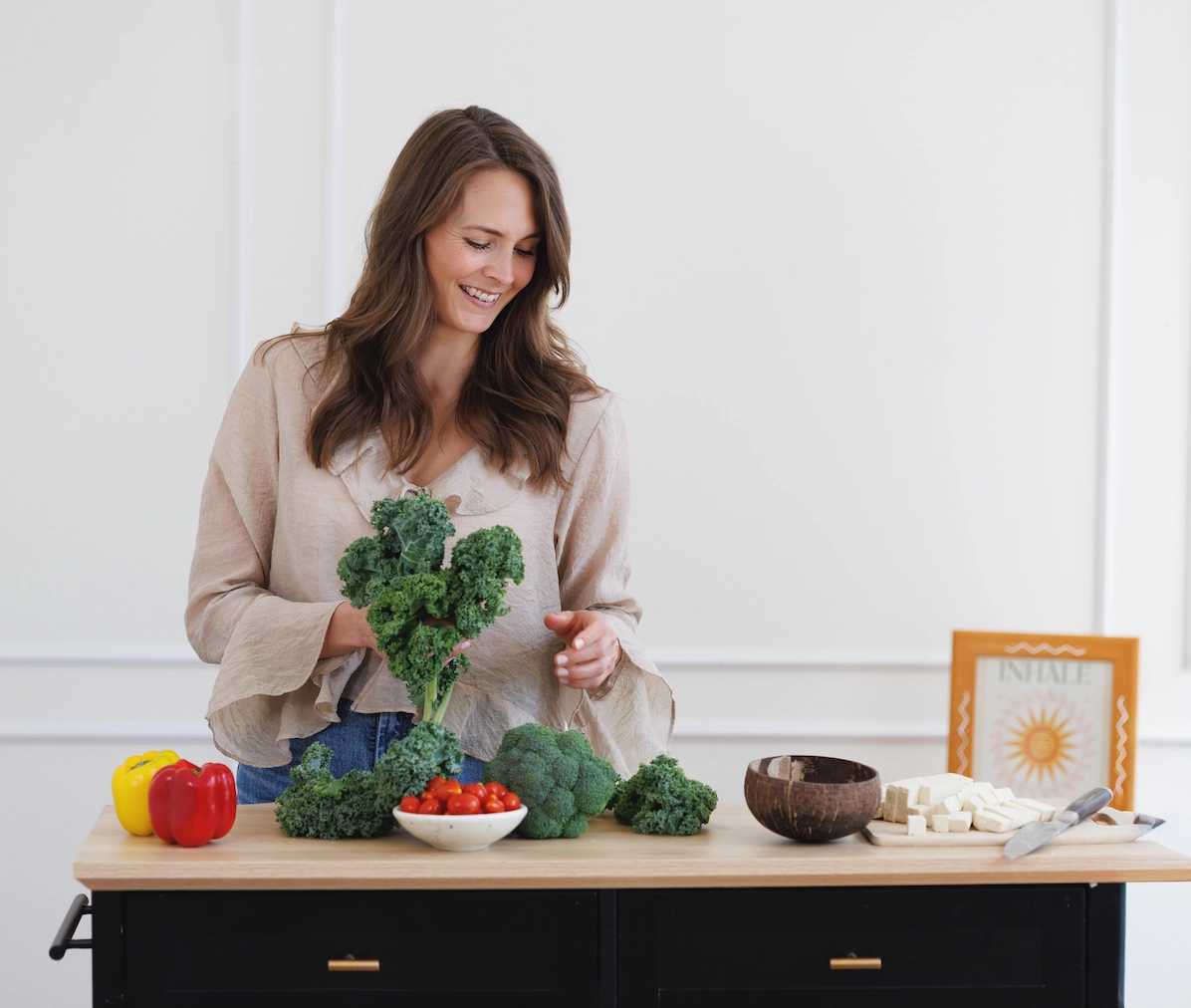 A vegan dietitian prepares vegetables like kale and broccoli at a kitchen island, with cherry tomatoes, red pepper, tofu, and a knife on the counter.