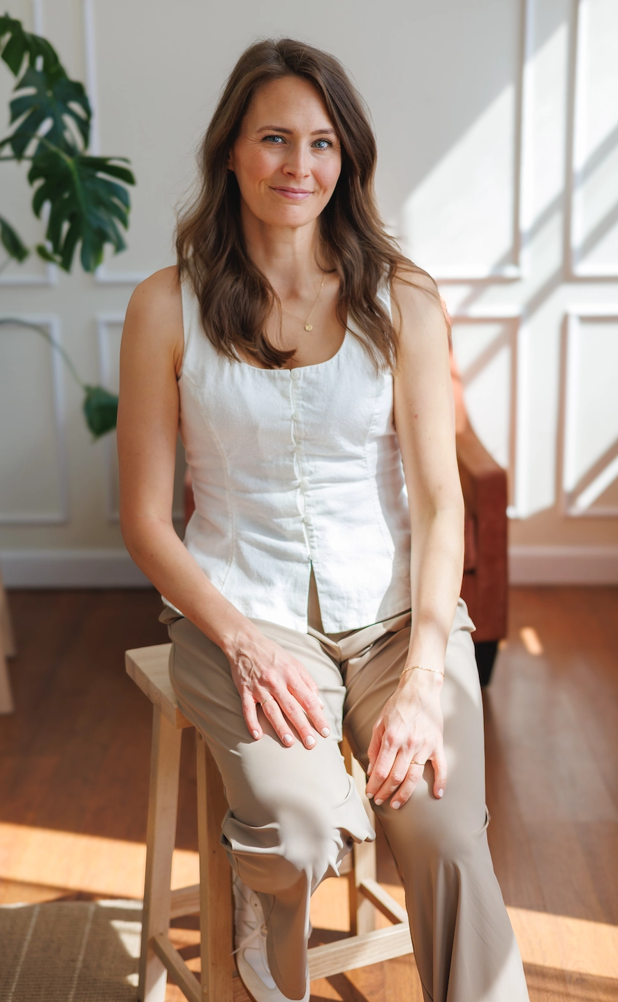 A woman with long brown hair, vegan dietitian Taylor Wolfram, wears a white sleeveless top and beige pants as she sits on a wooden stool indoors with sunlight streaming in.