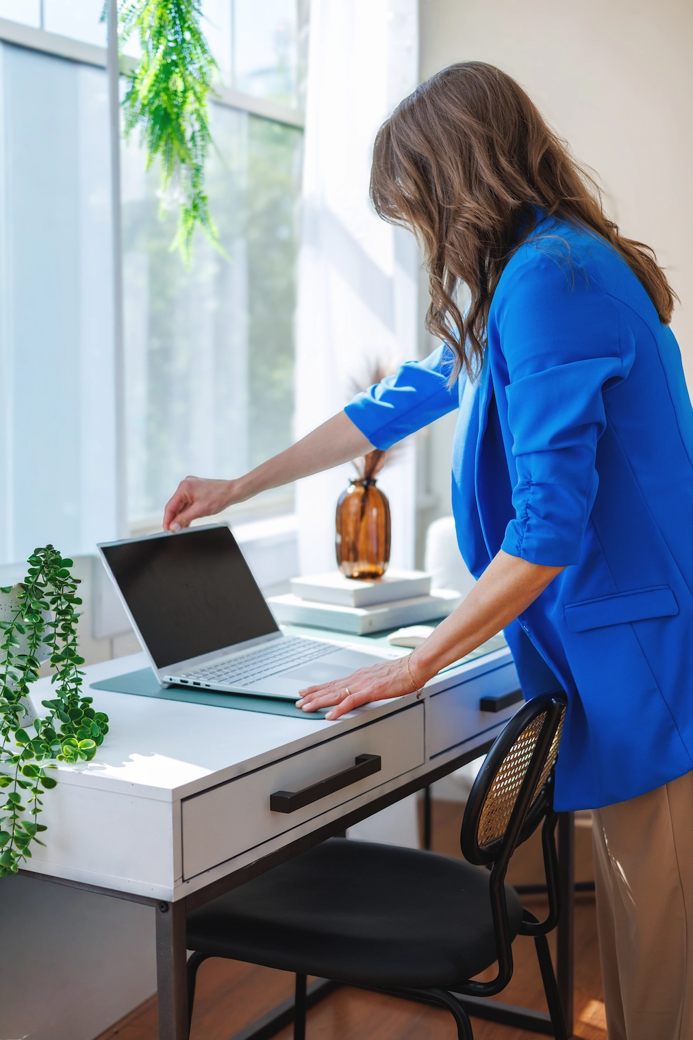 Dietitian Taylor Wolfram in a blue blazer opens her laptop on a white desk by a window, surrounded by plants and books.