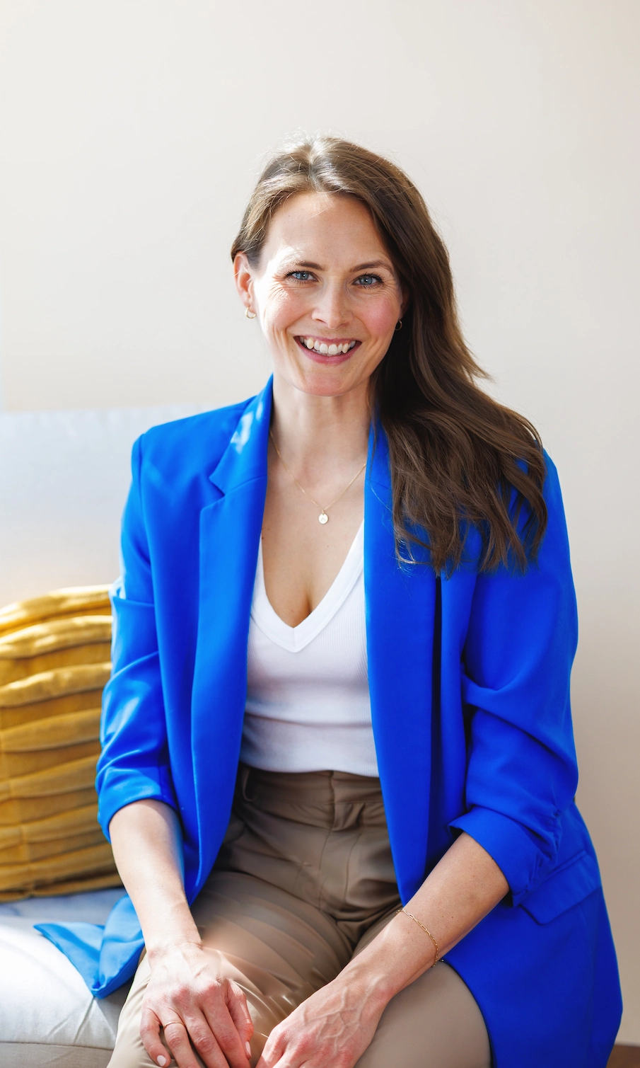 Chicago dietitian Taylor Wolfram, with long brown hair, wears a bright blue blazer, white top, and beige pants, sitting and smiling with a yellow pillow beside her.