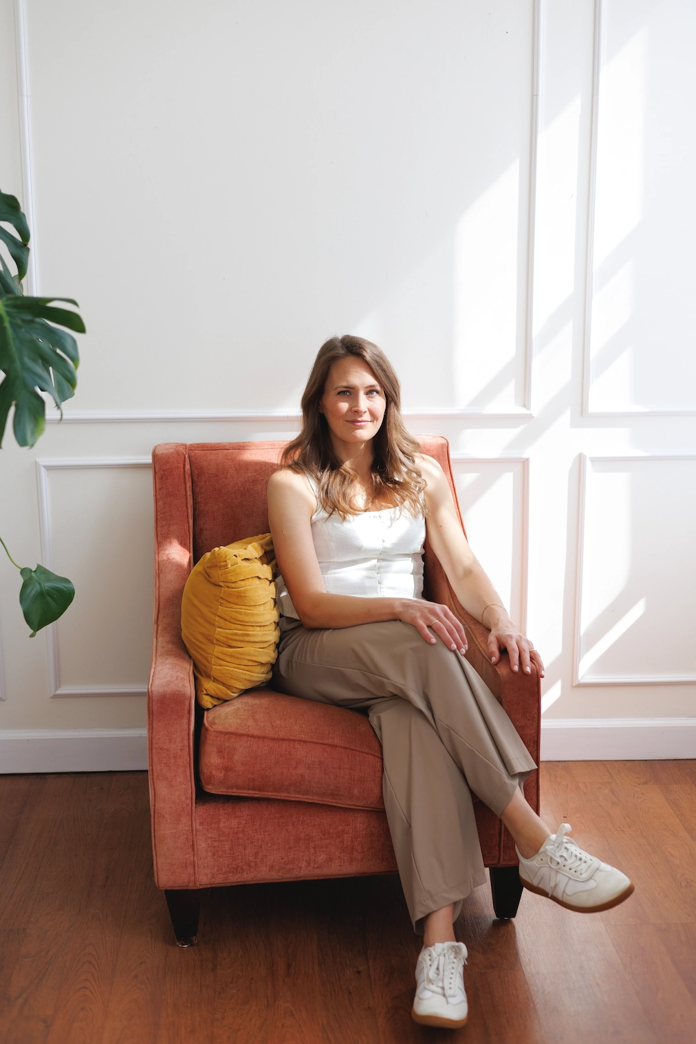 Woman dietitian sits on a pink armchair with a yellow pillow in a sunlit room, dressed in a white top, beige pants, and white sneakers.