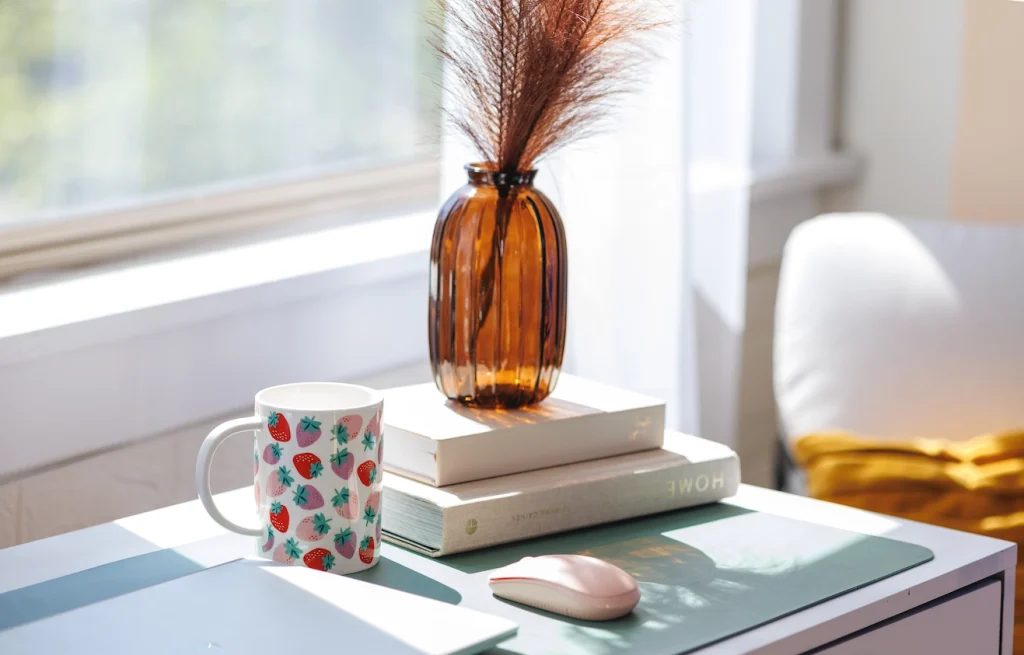 A dietitian’s desk with a strawberry-patterned mug, wireless mouse, two books, and dried grass in a vase, sunlight streaming through the window.