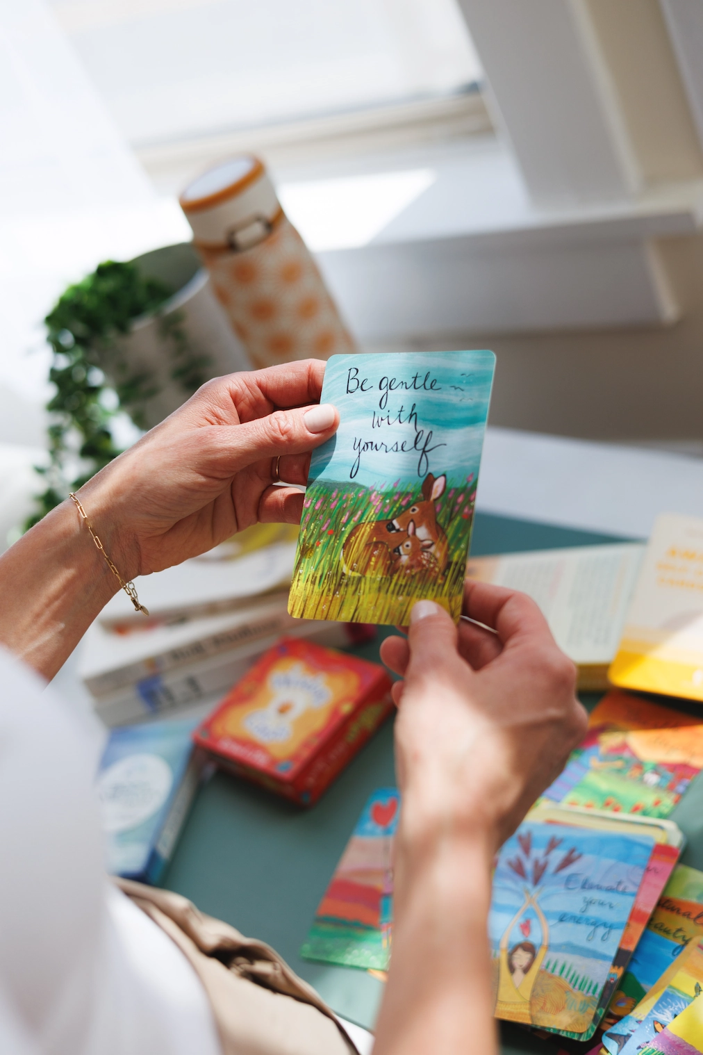 A person holds a card saying "Be gentle with yourself" above an illustrated deer; vegan dietitian Taylor Wolfram's colorful cards and books are on the table.