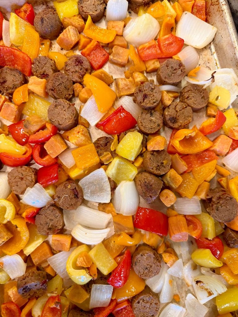 An overhead shot of a sheet pan containing round slices of vegan sausage and bite-size pieces of sweet potato, onion, and yellow, orange, and red bell peppers. 